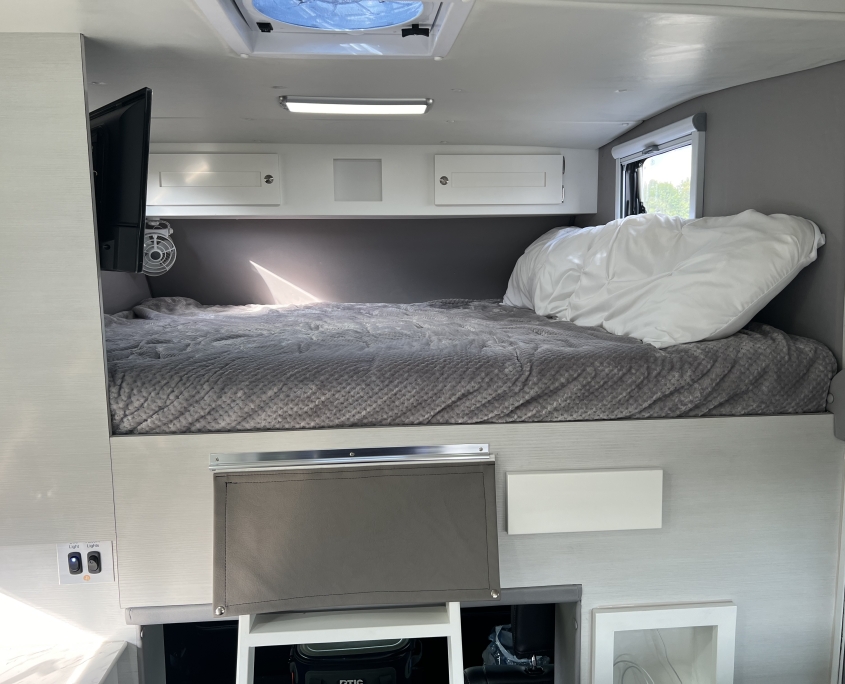 Panthera queen bed | Tiger Adventure Vehicles Tiger Panthera overhead cab-over bed with gray bedding, storage cabinets, and TV