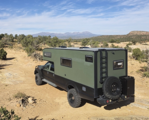 01_20241012_155023 1 EDITED | Tiger Adventure Vehicles | Tiger Adventure Vehicles Green off-road camper van parked in a dry desert landscape with sparse shrubs and distant mountains under a bright blue sky.