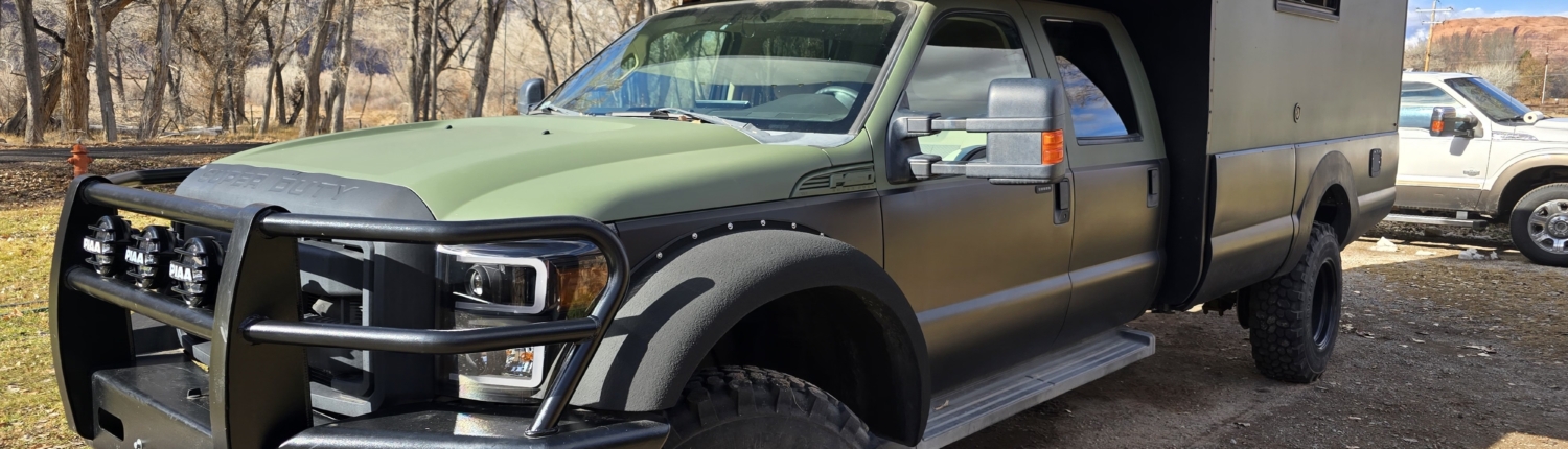 06_20260124_135320 edited | Tiger Adventure Vehicles Green pickup truck fitted with a large green overcab camper, parked on a dirt lot with leafless trees in the background.