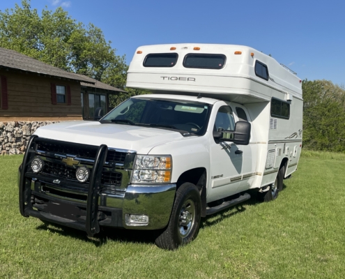 11aa695a-eca6-46d5-b8a6-f3c758596e5b | Tiger Adventure Vehicles | Tiger Adventure Vehicles White over-cab motorhome mounted on a pickup truck parked on a grassy lawn with a wooden house and trees in the background, under a blue sky.