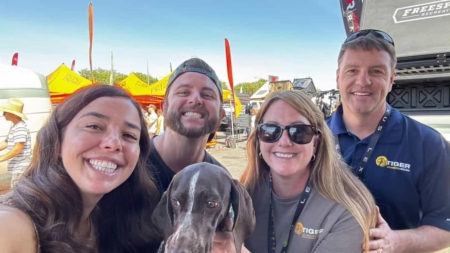 A group of four adults smiling at the camera with a gray hound in front, at an outdoor event with yellow tents in the background.