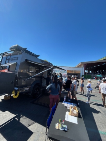 Line of people at an outdoor event beside a large gray expedition vehicle with a roof rack.