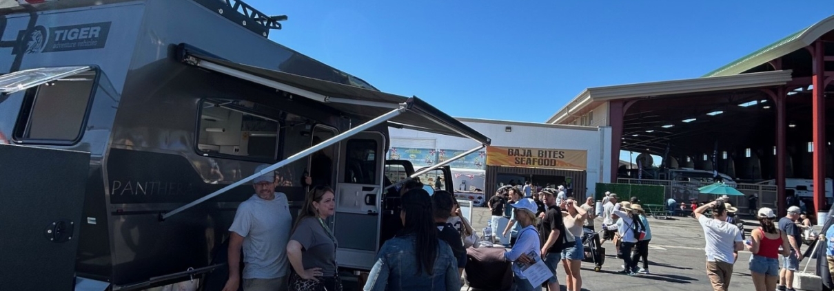 Outdoor festival scene with a gray RV/food truck and people queuing at booths under a bright blue sky; a table with brochures is in the foreground.