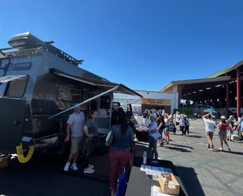 Outdoor festival scene with a gray RV/food truck and people queuing at booths under a bright blue sky; a table with brochures is in the foreground.