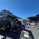 6f576bd2-1442-4d34-8491-0e7703384c13 | Tiger Adventure Vehicles Outdoor festival scene with a gray RV/food truck and people queuing at booths under a bright blue sky; a table with brochures is in the foreground.