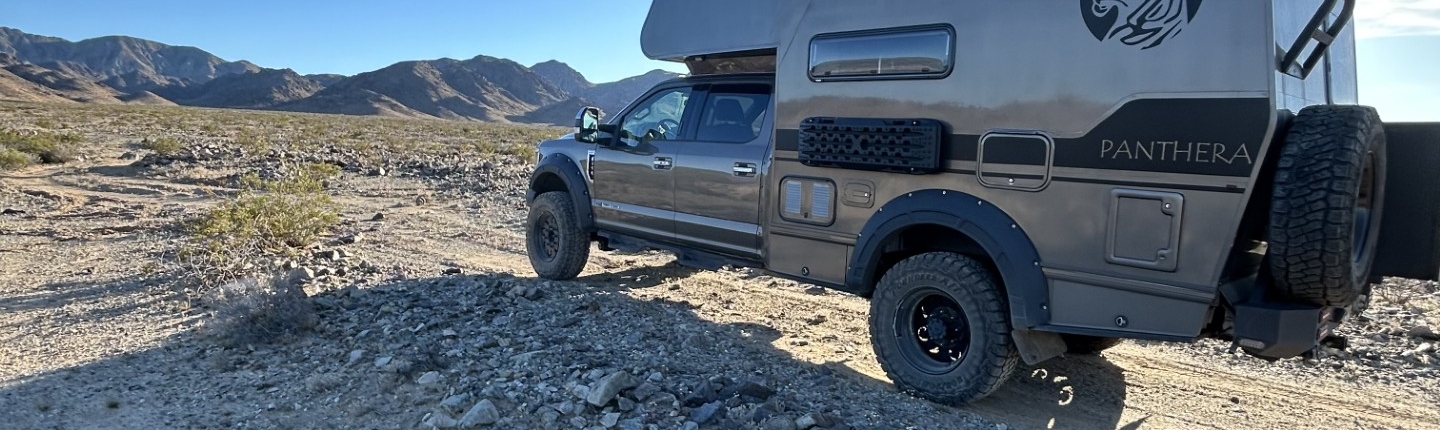 0241095c-43c7-45d5-b1c7-3c59bfdc0582 | Tiger Adventure Vehicles Camper-equipped pickup truck parked on rocky desert ground with distant mountains under a clear blue sky.