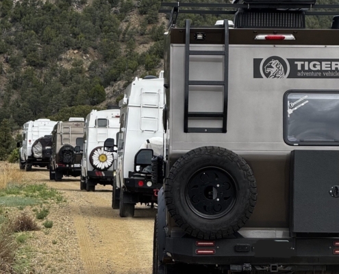 2ffad3ee-1302-4481-8e11-0063025eeb6b copy | Tiger Adventure Vehicles | Tiger Adventure Vehicles Line of overland campers parked on a dirt road with a forested hillside in the background; spare tire mounted on the rear of the lead vehicle.