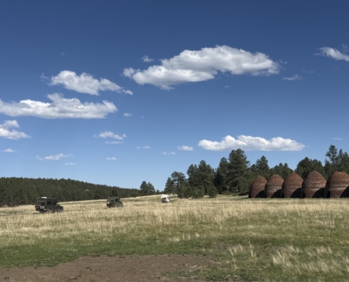 Wide grassy field with a few trucks on the left and a row of stacked round hay bales near the treeline under a clear blue sky.