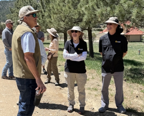 Group of adults wearing hats and outdoor clothing chat and smile beside a pine tree on a sunny day.