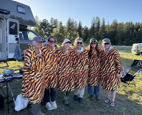 Six people wearing tiger-striped blankets pose together at a campsite with an RV and pine trees in the background.
