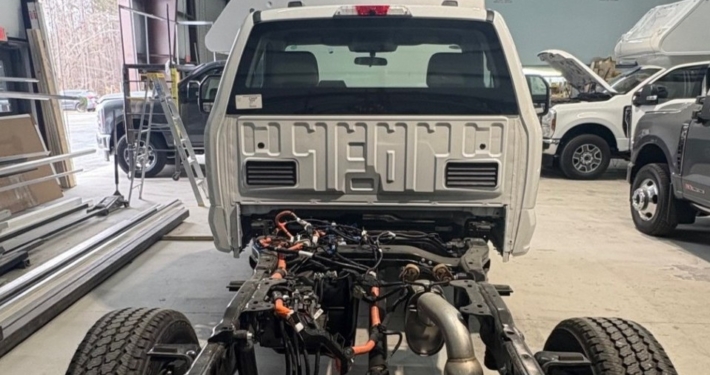 White pickup truck cab mounted on a bare chassis in a workshop, showing the exposed engine bay and drivetrain components for assembly/repair.