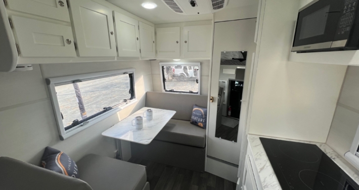 Bright white camper interior with upper cabinets, dinette table, gray seating, and a window with shade behind. Compact kitchen area with stove and sink nearby.