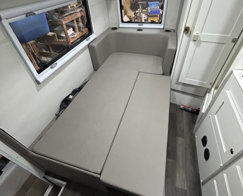 Compact RV interior with a gray L-shaped sofa bed, white overhead cabinets, and two windows showing a workshop outside the left and center walls; a door is on the right.