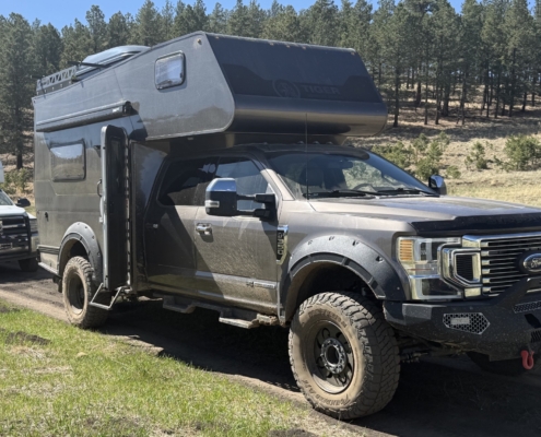 Grey Ford pickup with a large black rooftop camper attached, parked on a dirt trail with pine trees in the background on a sunny day.