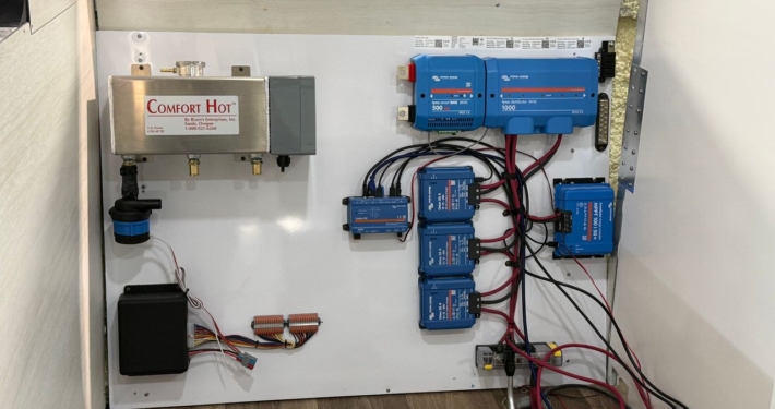 Electrical panel inside a trailer: blue inverters/charge controllers, red and black cables, and a silver Comfort Hot water box on the left.