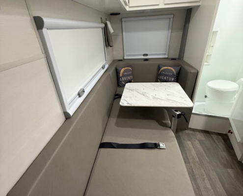 Compact camper interior with beige L‑shaped seating, a marble tabletop, and white overhead cabinets; window shade closed and a toilet visible at right.