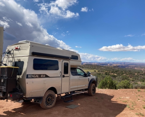 White Bengal 4x4 camper van parked on a dirt overlook, desert canyon and blue sky with clouds in the distance.