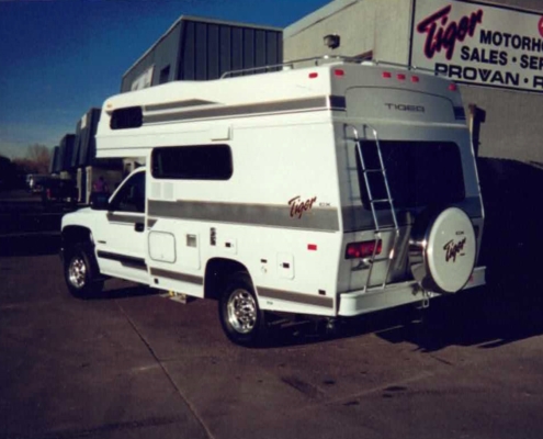 White Tiger motorhome with side stripe and rear ladder parked in a dealership lot beside a signs reading Tiger Motorhomes.