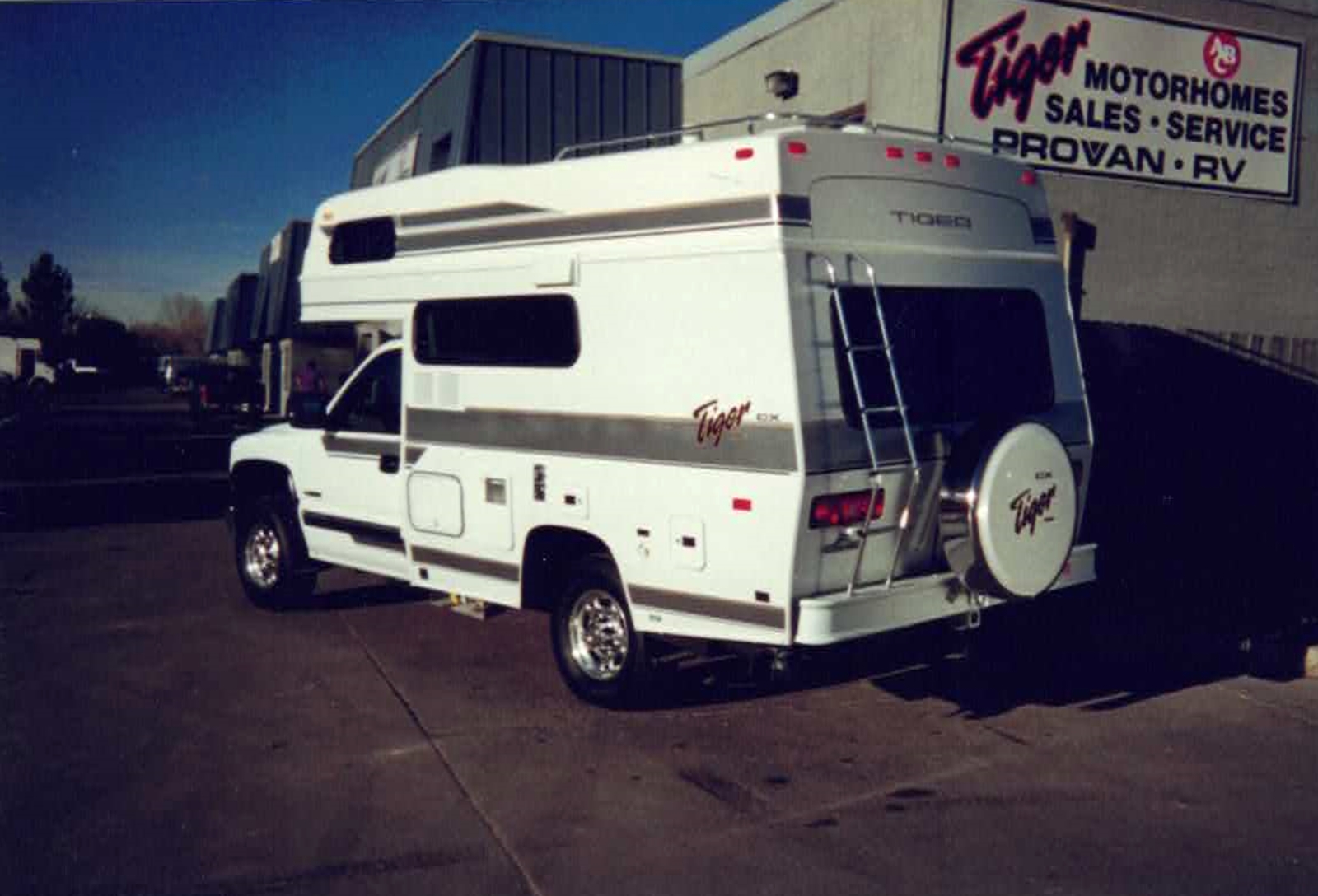 White Tiger motorhome with side stripe and rear ladder parked in a dealership lot beside a signs reading Tiger Motorhomes.