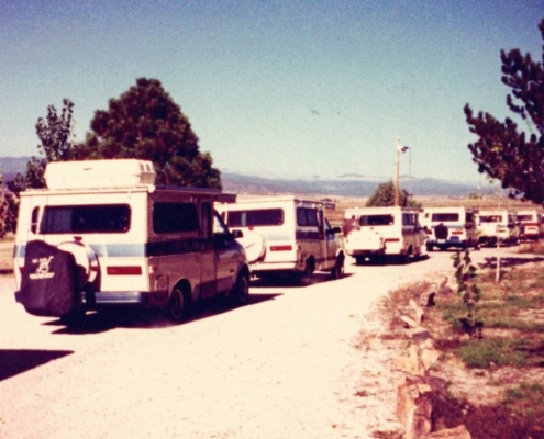 Line of white and blue camper vans parked along a dusty road with trees and hills in the background.