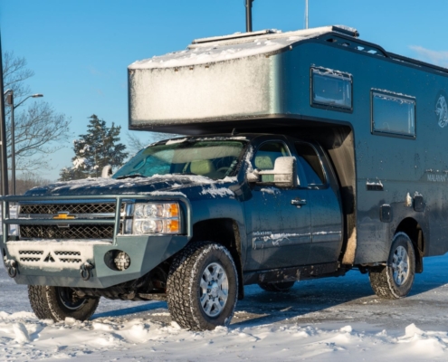 Blue pickup with a large overcab camper shell parked in a snowy lot, with snow on the hood and roof keeping the scene wintry and rugged.