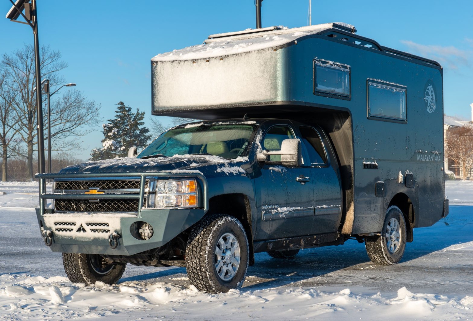 Blue pickup with a large overcab camper shell parked in a snowy lot, with snow on the hood and roof keeping the scene wintry and rugged.