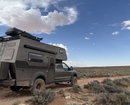 Overland camper truck labeled Panthera, with a rooftop rack and spare tire, parked on a dusty desert trail under a blue sky with clouds.