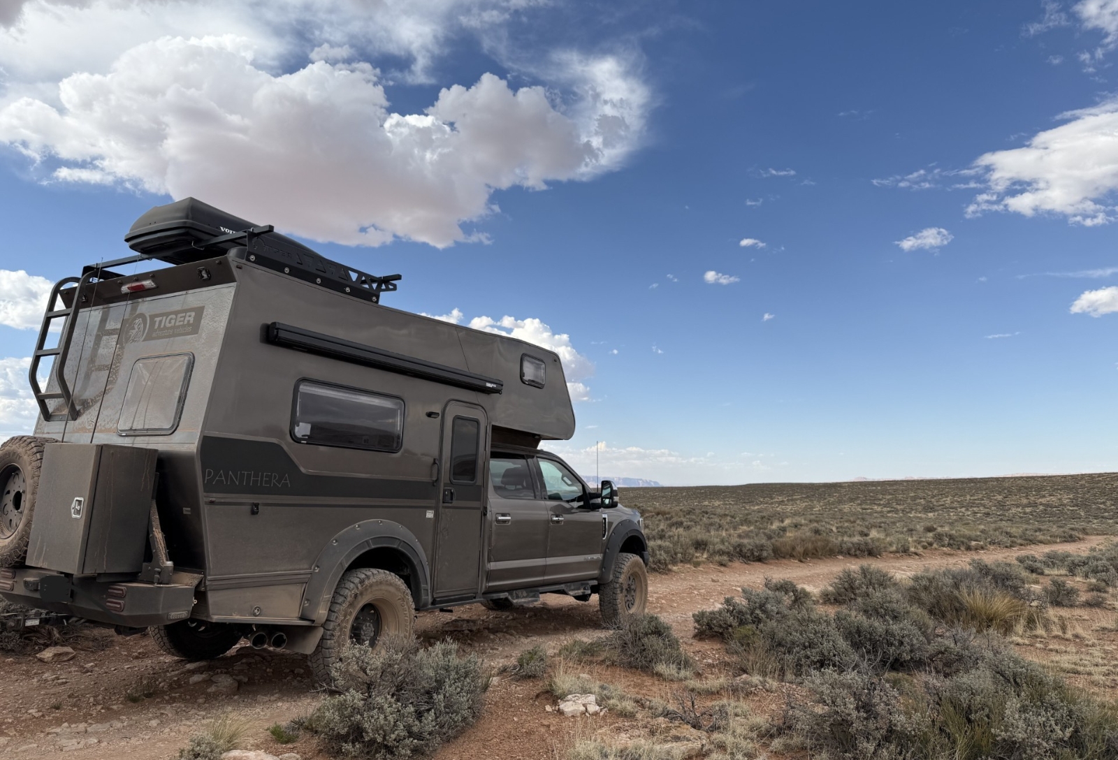 Overland camper truck labeled Panthera, with a rooftop rack and spare tire, parked on a dusty desert trail under a blue sky with clouds.