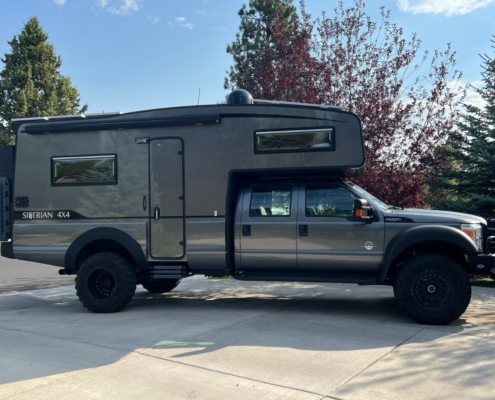 Grey pickup-based camper van with a raised living quarters over the cab, parked on a driveway with trees in the background