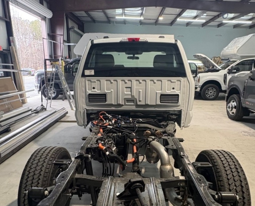 White truck cab mounted on a bare chassis in a workshop, with exposed engine bay, wiring, and components visible; other vehicles nearby in the shop