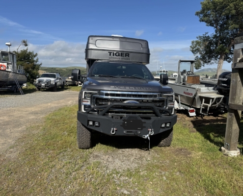 IMG_1671 | Tiger Adventure Vehicles | Tiger Adventure Vehicles Front view of a dark gray pickup truck with a large silver rooftop cargo box labeled 'TIGER adventure vehicles' in a boatyard.