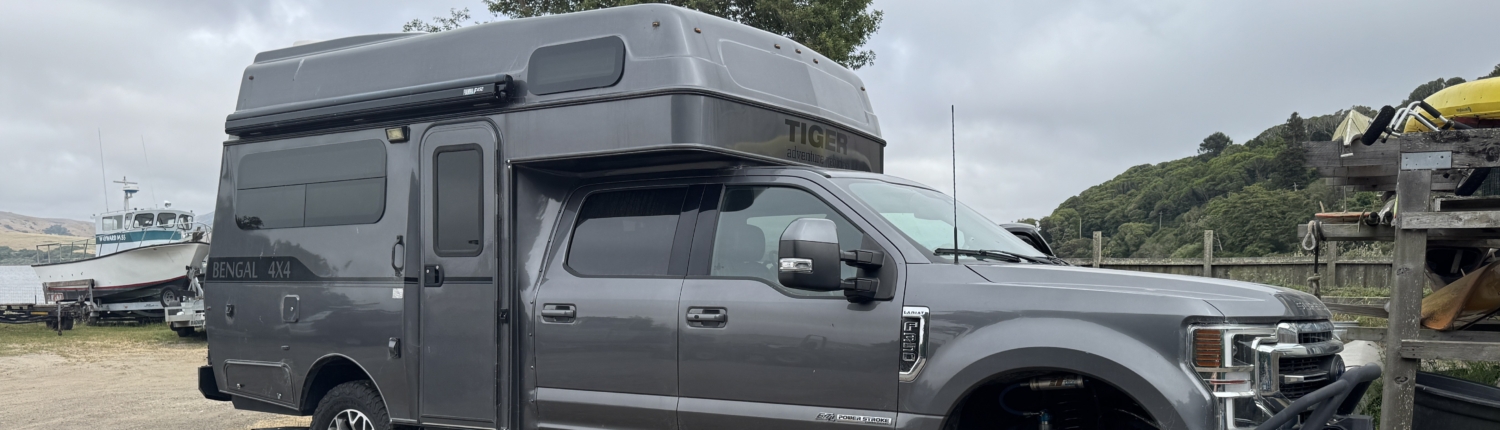 Gray camper van with a raised roof and off-road tires parked on a grassy area, boats in the background and cloudy sky above.