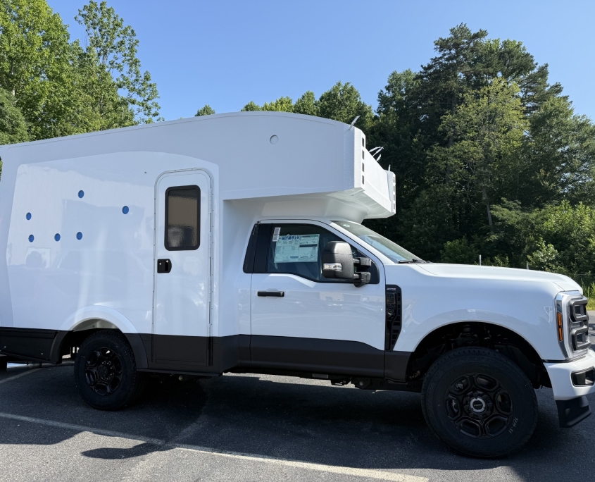 White camper attached to a pickup truck with an elevated living area, parked on asphalt with green trees and blue sky behind.