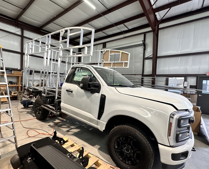 White pickup truck on a flatbed trailer inside a workshop, with a tall metal frame structure being built on the trailer.