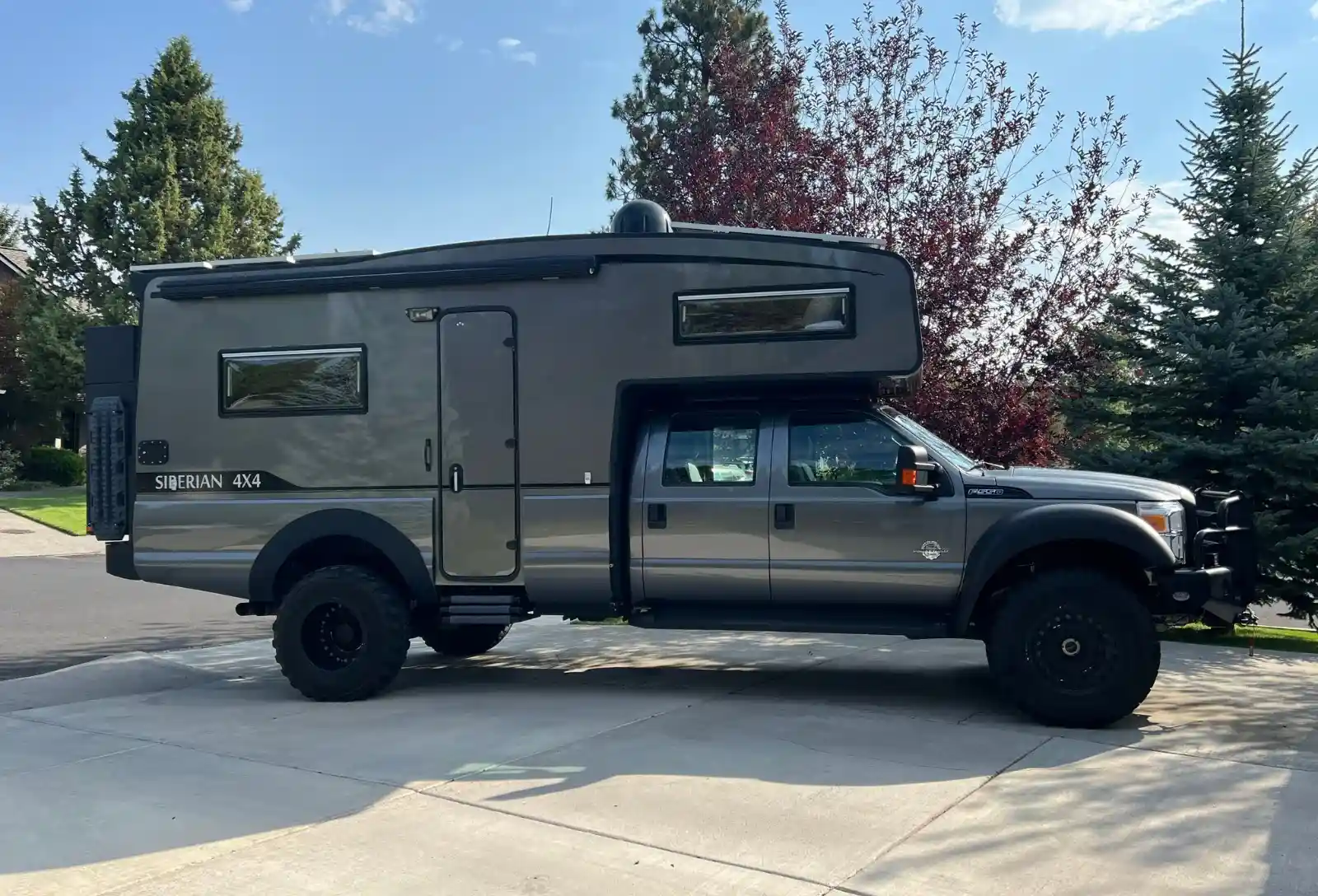 tiger-Siberian-Model | Tiger Adventure Vehicles Grey pickup-based camper van with a raised living quarters over the cab, parked on a driveway with trees in the background