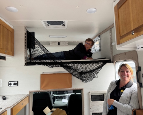 Woman lying on the upper bunk of a camper with a safety net, smiling; another woman stands near the doorway inside the RV.