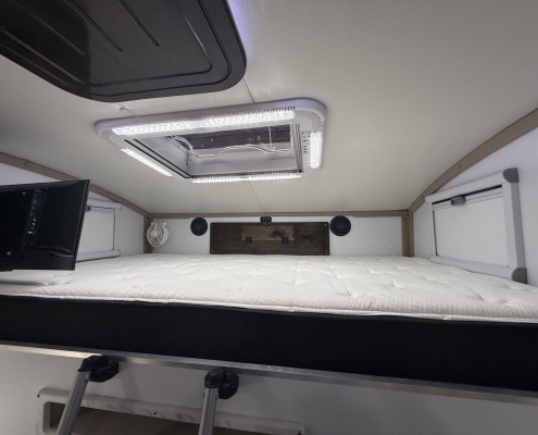Interior of a camper van showing a bunk with a white mattress and an illuminated ceiling hatch/vent, metal ladder below, and exposed wood accents.