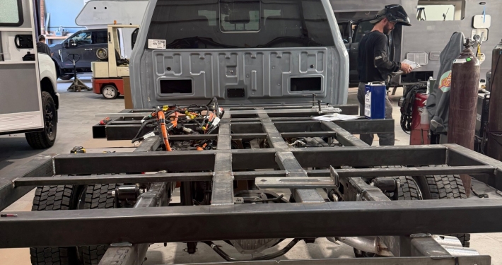 | Tiger Adventure Vehicles Workshop scene with a metal vehicle chassis framework in the foreground and a person in a shop working near a gray pickup bed in the background
