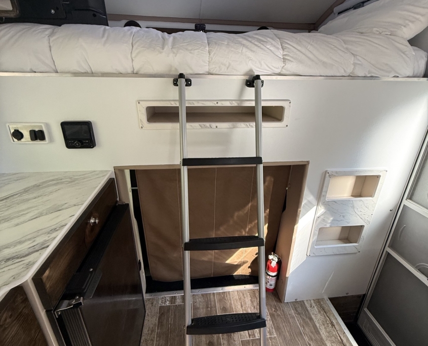 Interior of a camper van with a top bunk and metal ladder, kitchenette on the left and a skylight above the bed area