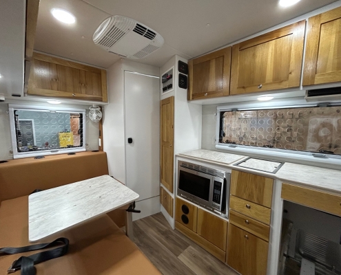 Interior of a wood-cabinet RV kitchen with table and brown bench seating, microwave and oven, and window panels on the right side.