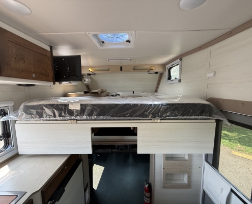 Interior of a camper van showing a bed platform with protective plastic, white cabinets, and a small kitchen counter area to the left