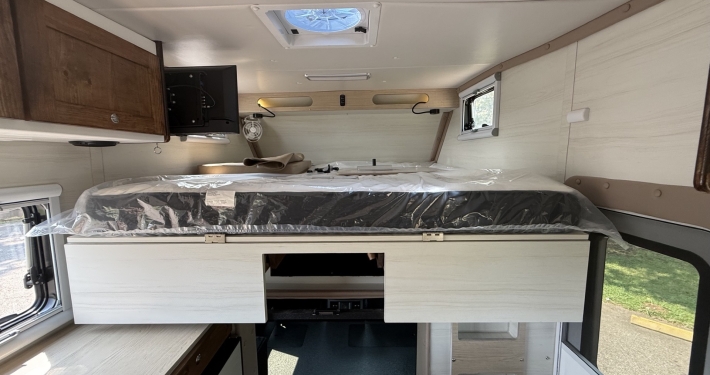 Overhead King Bed (north-south) | Tiger Adventure Vehicles Interior of a camper van showing a bed platform with protective plastic, white cabinets, and a small kitchen counter area to the left