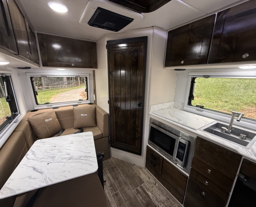 Interior view of a compact RV kitchen and dining area with dark wood cabinets, beige sofa, marble tabletop, and a windowed counter space on the right.