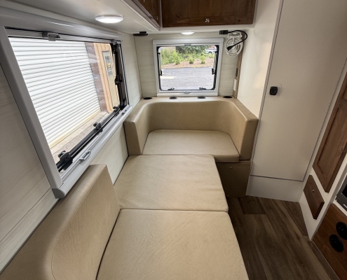 Interior of a compact camper with a beige L-shaped sofa, wooden overhead cabinets, and a windowed seating area facing an outside view