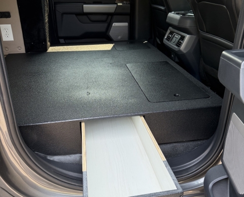 Interior of a vehicle with a raised, black textured sleeping/storage platform and a slide-out drawer housing a circular light, with front seats visible