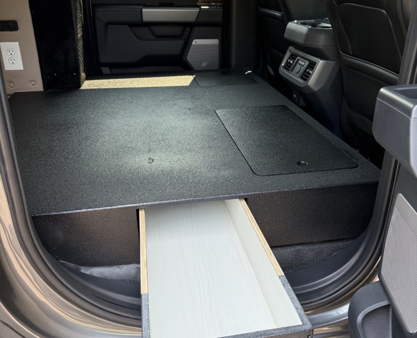 Flat platform with drawers and hatches | Tiger Adventure Vehicles Interior of a vehicle with a raised, black textured sleeping/storage platform and a slide-out drawer housing a circular light, with front seats visible