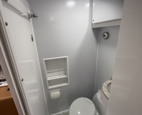 Compact white bathroom with a toilet, small vanity sink, and wall-mounted mirror cabinet in a narrow space. A towel bar, recessed shelf, and ceiling light are visible; a floor drain is near the shower area.