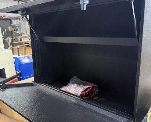 Open black metal safe with lid raised; empty interior and a bag of red cables resting on the bottom, a person’s hand at the front edge.