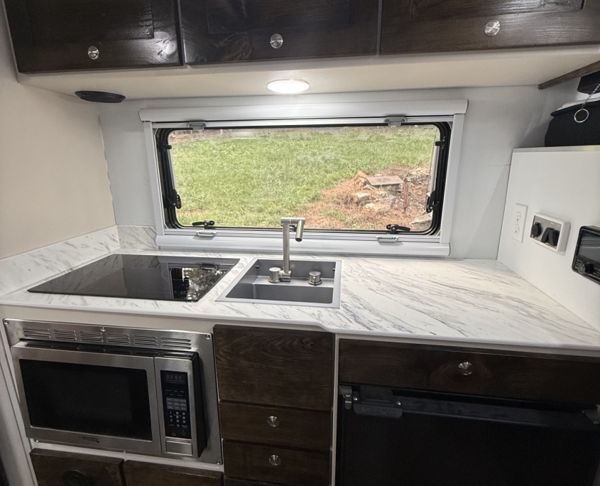 RV kitchen with dark wood upper cabinets, marble countertops, and a window over the sink.