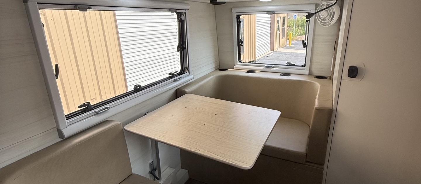 Dinette | Tiger Adventure Vehicles Interior of a small RV dining area with beige cushioned bench seats around a light wood table, dark wood overhead cabinets, and two windows with blinds letting in daylight.